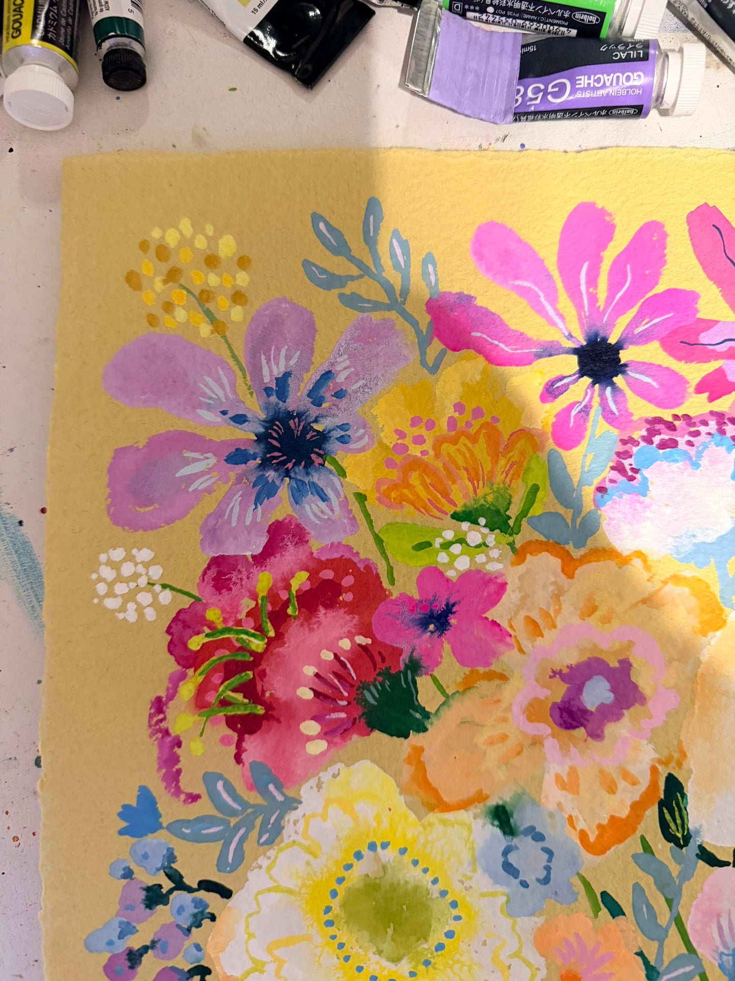 Colorful floral painting on a yellow background with art supplies in the background.