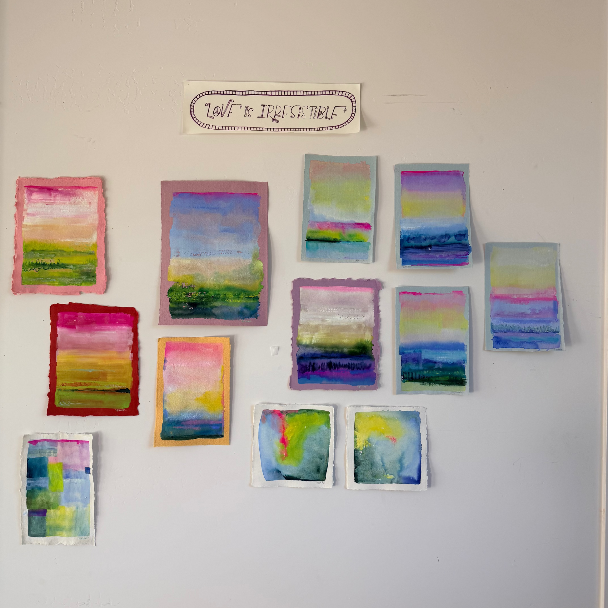 Colorful abstract paintings on a white wall with a sign above them.